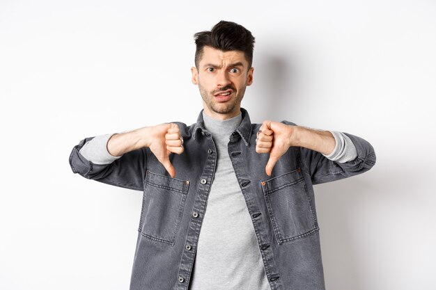 Disappointed guy showing thumbs down and frowning, looking skeptical at something bad, dislike and disapprove awful thing, standing on white background.