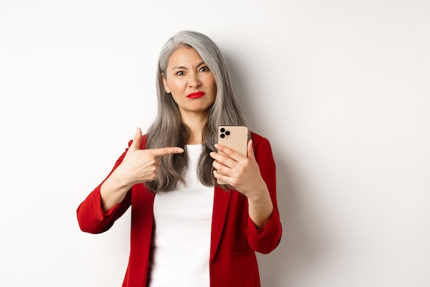 Disappointed mature asian woman complaining, pointing finger at smartphone and looking displeased, standing over white background