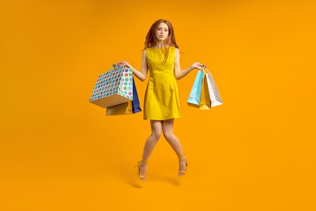 Disappointed red haired woman in dress holds shopping packages, upset by shopping. Beautiful female looks unhappy, dislikes purchase. Bored and tired woman buys presents, isolated on yellow, jumping