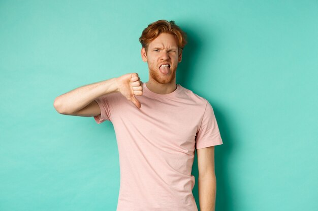 Photo disappointed redhead man showing thumbs-down and tongue, epxress dislike and disapproval, standing over turquoise background