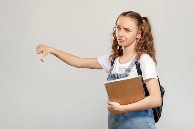 失望した学生の肖像画試験の失敗ライトコピースペースの背景に分離された親指を下に向けた拒否ジェスチャーを示す本を持つ懐疑的な女の子間違った選択悪い考え恥の敗者