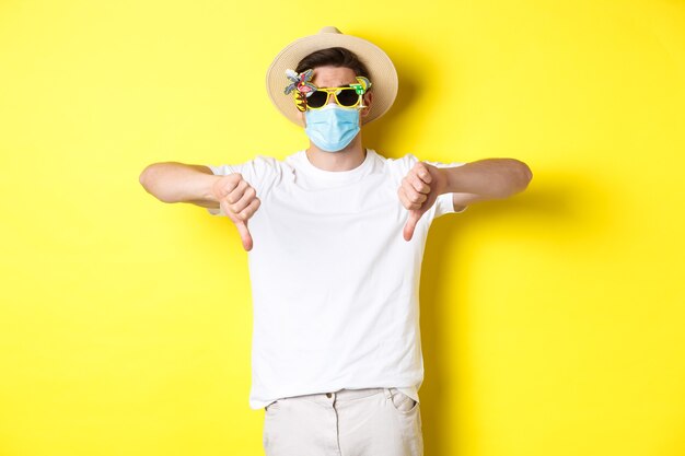 Disappointed tourist complaining on lockdown during pandemic, wearing medical mask and sunglasses, showing thumbs down.