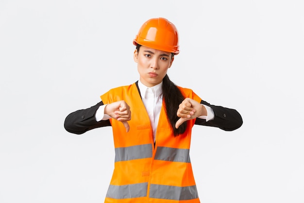 Disappointed and upset asian female chief engineer inspect bad work, showing thumbs-down disappointed, wear safety helmet and complaining weak awful work of architect, white background