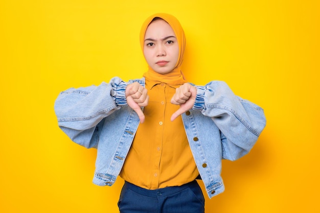 Disappointed young Asian woman in jeans jacket showing thumb down with expressing disapproval isolated over yellow background