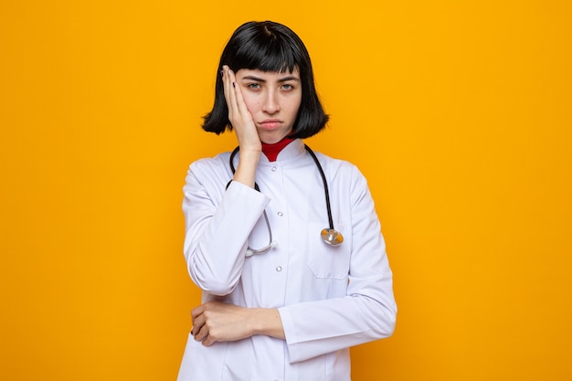 Разочарованная молодая симпатичная кавказская девушка в медицинской форме со стетоскопом кладет руку ей на лицо и смотрит вперед