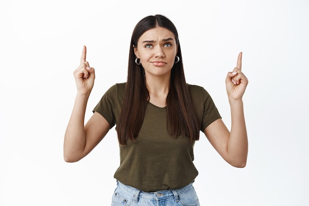 Disappointed young woman with skeptical grimace pointing fingers up looking at something upsetting with dislike standing in casual tshirt against white background