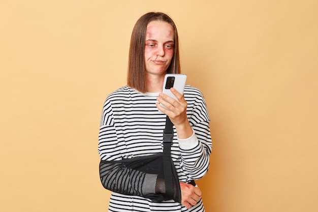 Disappointedinjuredwoman with bruise on face wearing bandage using mobile phone isolated over beige background browsing internet needs psychological help after violence