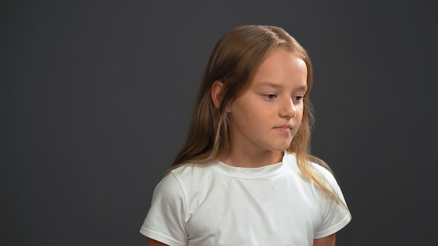 Disappointment little girl with long blond hair, looking thoughtful down wearing white t-shirt and black pants isolated on black wall
