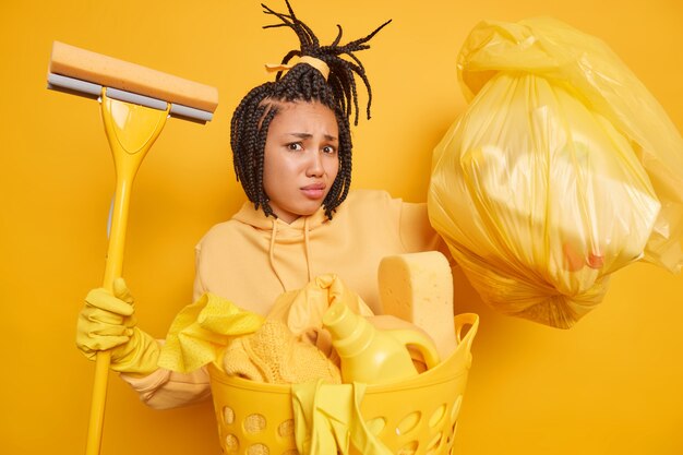 Discontent African American woman has dreadlocks busy doing housework holds bag with detergents