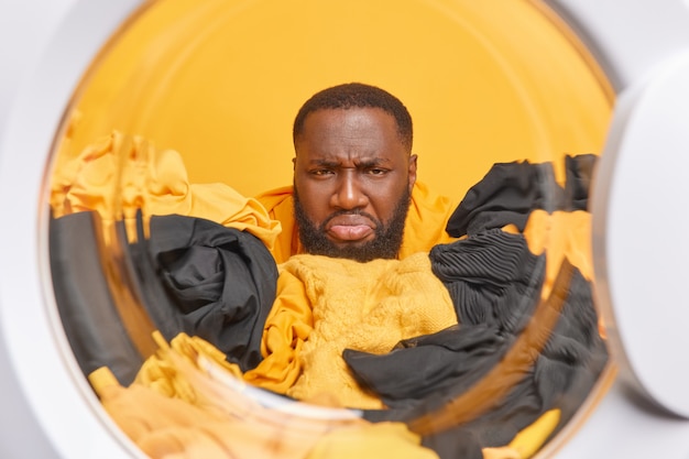 Discontent bearded male householder does washing poses through washing machine drum does housework has unhappy face expression as doesnt want to do laundering