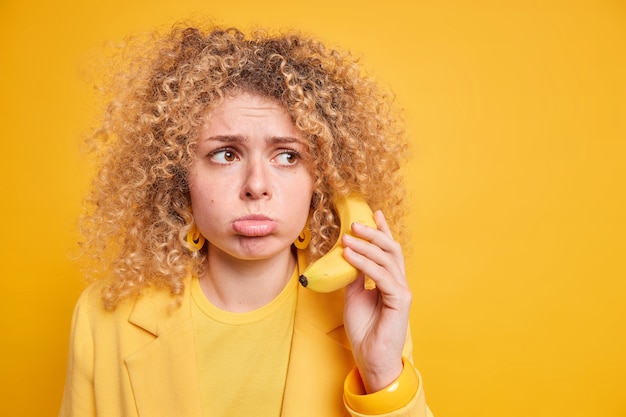 Discontent curly haired woman purses lips looks sadly away holds banana near ear waits for call from boyfriend has dejected face expression wears elegant clothes isolated over yellow wall