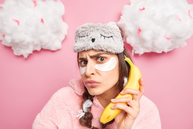 Фото Недовольная женщина сердито смотрит в камеру, накладывает коллагеновые пластыри под глаза, держит банан как телефон