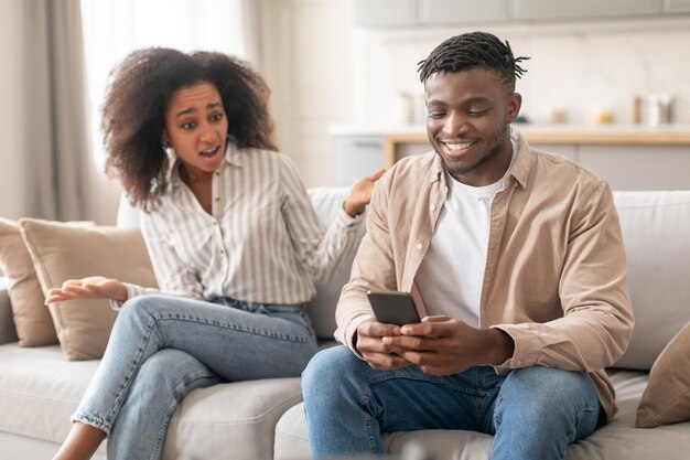 Photo discontented black wife looking at husband browsing on phone indoor