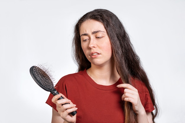 A discontented brunette woman holds a comb with a bunch of torn hair and looks at her hair