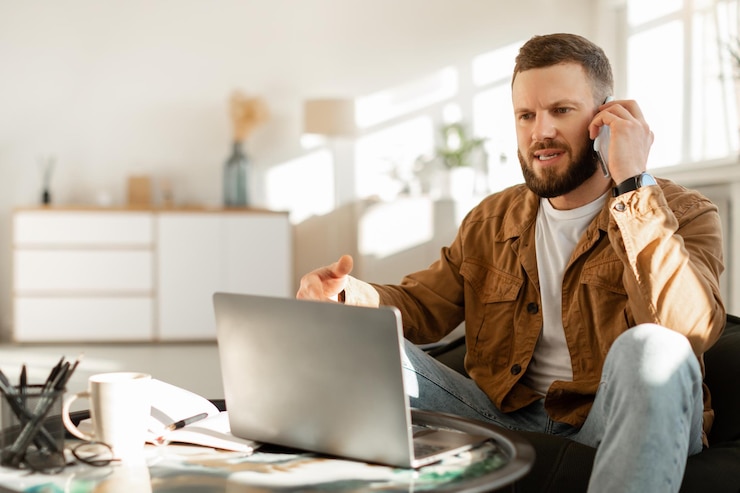 Photo discontented male freelancer talking on cellphone using laptop at home