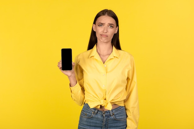 Discontented millennial woman showing cellphone with blank screen yellow background