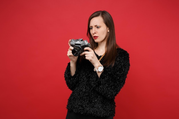 Discouraged young woman in black fur sweater holding, looking on retro vintage photo camera isolated on bright red wall background. People sincere emotions, lifestyle concept. Mock up copy space.