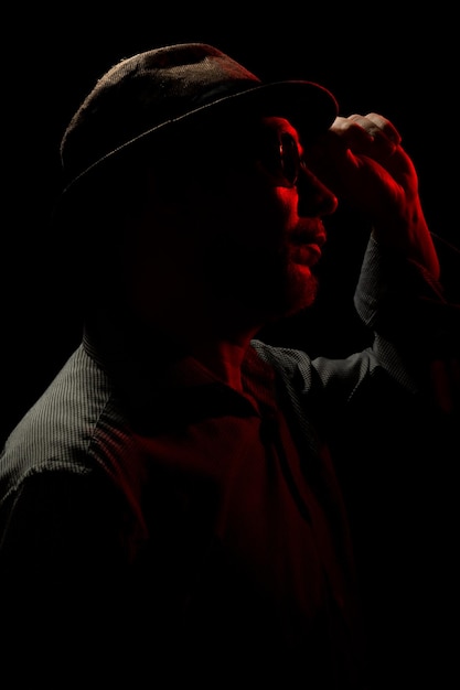 Discreet portrait of man wearing hat and sunglasses in darkness with red light on face making light hand gestures Dark room art