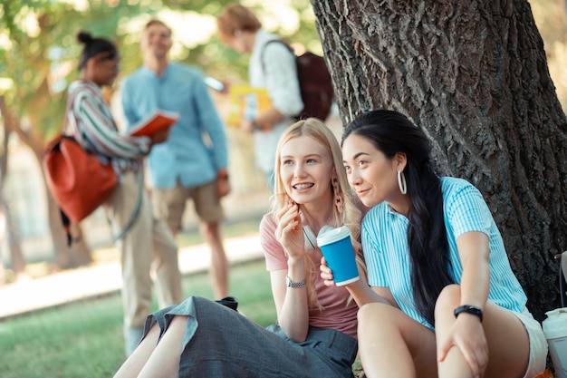 Discussiëren over uiterlijkheden. Twee lachende meisjes zitten onder een grote boom koffie te drinken en voorin te praten over hun groepsgenoten.