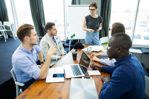 Discussing new business ideas. Cheerful young woman standing near whiteboard and smiling while her colleagues sitting at the desk.