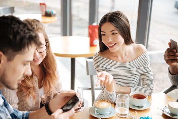 Discussing social media. Young smiling asian girl is gesting to her friends at cafe using smartphones.