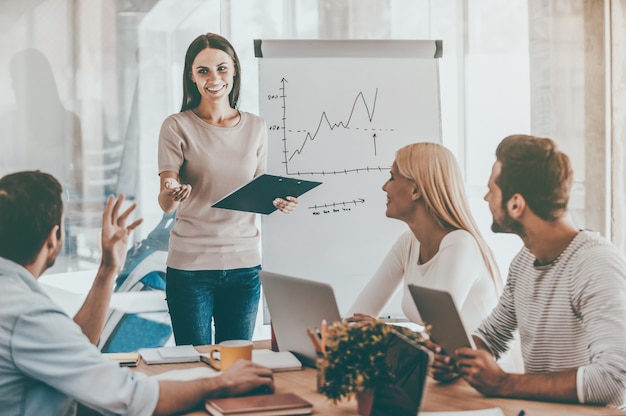 Discussing some business issues. Cheerful young woman standing near whiteboard and smiling while her colleagues sitting at the desk