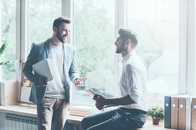 Discussing their successful project. Two young businessmen in smart casual wear talking and gesturing while leaning at the window sill in office