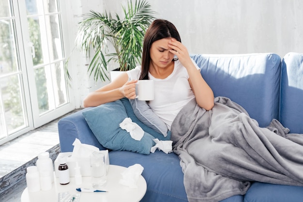 Diseased female lying on sofa with a cup of tea while touching her head