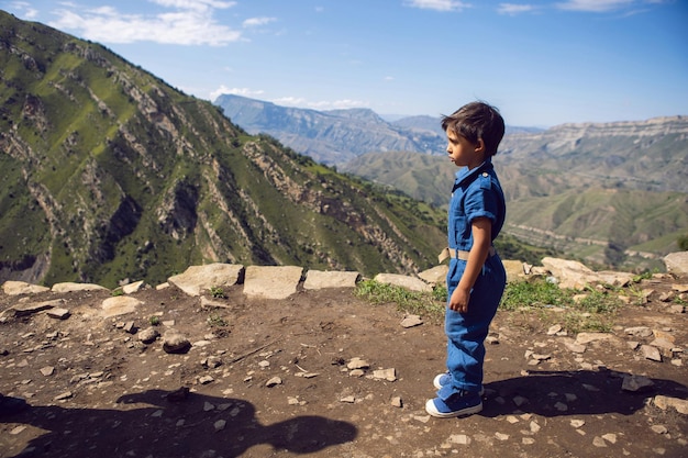 Disgruntled child is a traveler boy in blue jumpsuit standing in the mountains in summer
