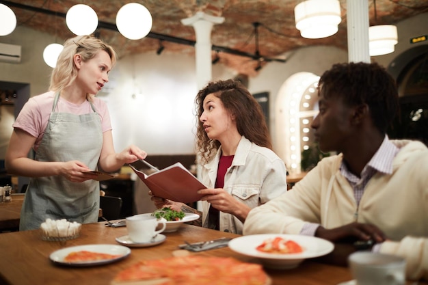 Disgruntled curlyhaired young female restaurant guest sitting at table with boyfriend and reading menu while talking to waitress