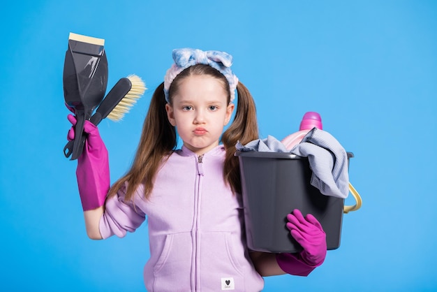 Disgruntled grumpy angry little girl in two ponytails with headband holds cleaning accessories