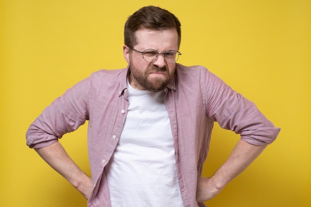 Disgruntled man in glasses and casual clothes stands akimbo and looks judgmental and vicious