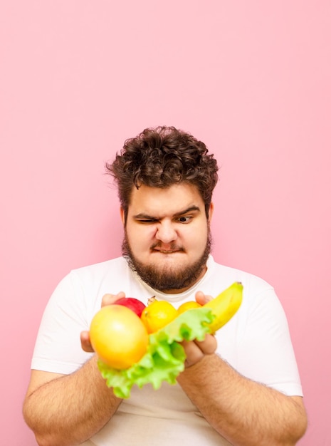 disgruntled young fat man with fruits in his hands on a pink background looking at raw food