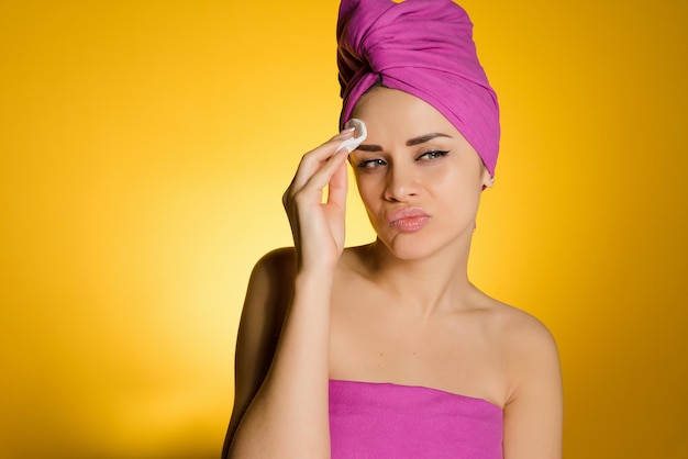 Disgruntled young girl with a pink towel on her head cleans the skin with a cotton swab