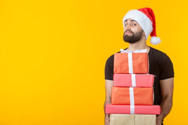 Disgruntled young man with a beard in a Santa Claus hat holds five gift boxes posing on a yellow wall with copyspace. Concept of gifts and greetings for Christmas 