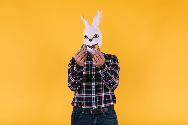 Photo disguised person in rabbit mask wearing checkered shirt and jeans holding easter eggs on yellow background carnival party easter and celebration concept