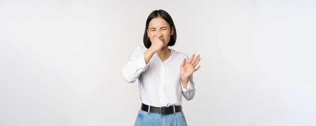 Азиатская девушка с отвращением закрывает нос и показывает жест отказа, отвратительный неприятный запах, стоящий на белом фоне