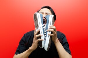 Disgusted asian man with black t-shirt holding a pair of stinky shoes isolated on red background.