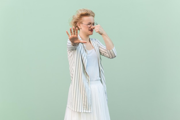 Disgusted blonde woman wearing striped shirt and skirt pinching her nose showing stop gesture