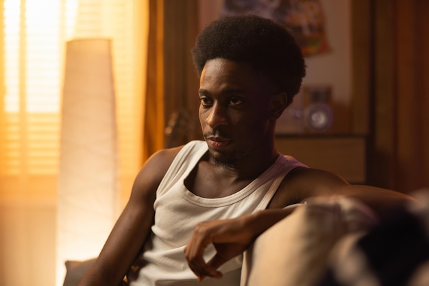 A disgusted man with a strange face sits on the sofa in a comfortable sleeveless white tshirt