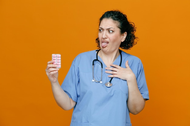 Фото Отвратительная женщина-врач средних лет в униформе и со стетоскопом на шее держит упаковку капсул, глядя на нее, держа руку на груди, показывая язык изолирован на оранжевом фоне