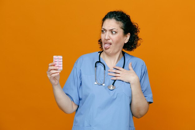 Disgusted middleaged female doctor wearing uniform and stethoscope around her neck holding pack of capsules looking at it while keeping hand on chest showing tongue isolated on orange background