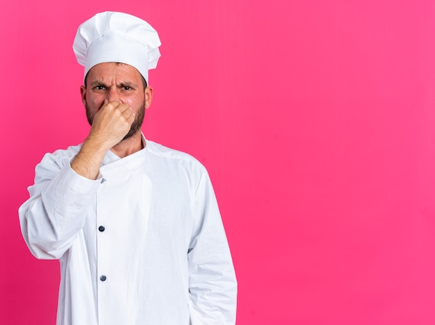 역겨운 젊은 백인 남성 요리사 유니폼을 입고 모자를 쓰고 복사 공간이 있는 분홍색 벽에 격리된 나쁜 냄새 제스처를 하는 카메라를 바라보고 있습니다.