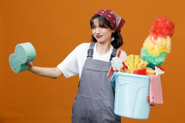 Disgusted young female cleaner wearing uniform and bandana holding bucket of cleaning tools stretching sponge out looking at side isolated on orange background