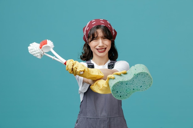 Disgusted young female cleaner wearing uniform bandana and rubber gloves stretching toilet brushes and sponge out towards camera looking at camera showing no gesture isolated on blue background