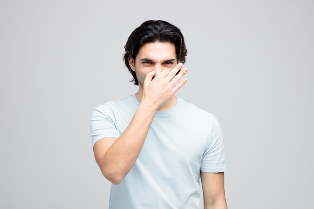Disgustato giovane bell'uomo che tiene il naso che mostra un gesto di cattivo odore mentre guarda la fotocamera isolata su sfondo bianco