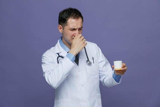 Disgusted young male doctor wearing medical robe and stethoscope around neck holding tablet container looking at it making bad smell gesture isolated on purple background