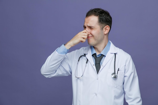 Disgusted young male doctor wearing medical robe and stethoscope around neck looking at side making bad smell gesture isolated on purple background