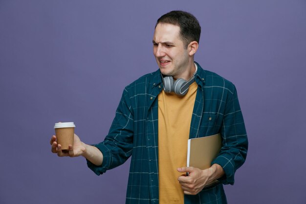 Disgusted young male student wearing headphones around neck holding note book under arm holding paper coffee cup looking at it isolated on purple background