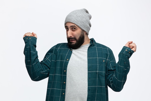 Disgusted young man wearing winter hat keeping fists in air looking at camera isolated on white background